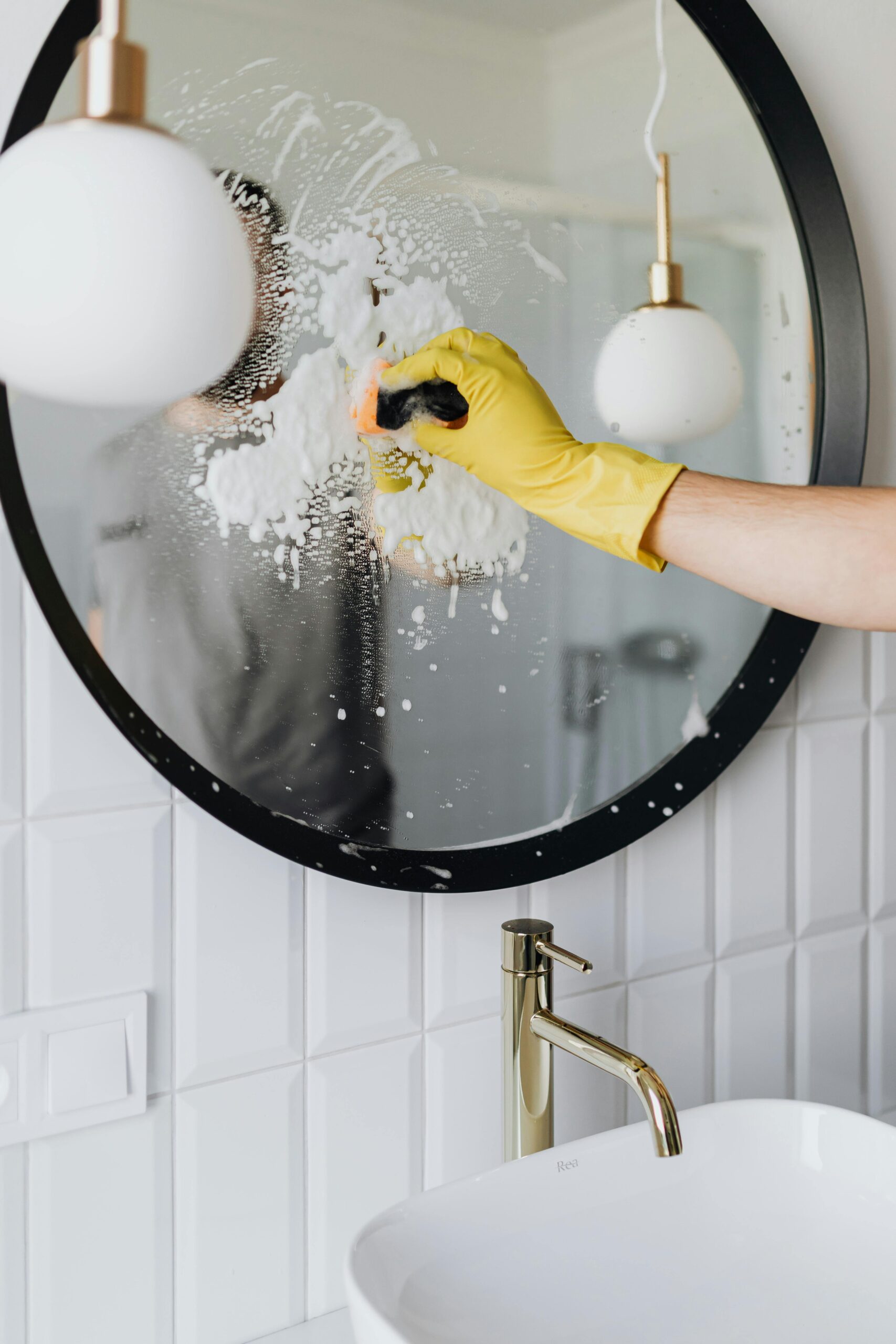 A person wearing a yellow glove cleans a bathroom mirror with foam spray.
